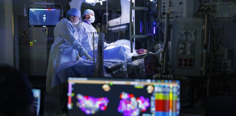 two baptist health heart surgeons performing a procedure in a dark operating room with lighting focused on patient, along with a colorful computer screen in the foreground.